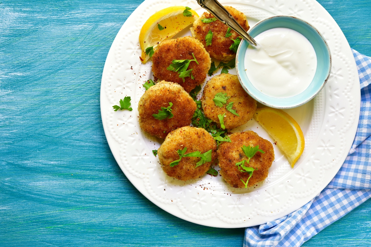 Ricette con merluzzo: idee facili, veloci e leggere 1 piatto tondo su base azzurra con polpette di merluzzo al forno e ciotola con salsa bianca