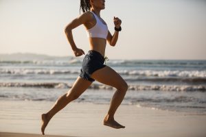 Home Page 69 donna che ha scelto come sport anticellulite la corsa. Corre con gli shorts e top bianco sulla spiaggia in riva al mare piedi nudi