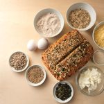 Pane proteico: benefici, ingredienti e la ricetta da fare in casa 1 In primo piano su uno sfondo un pane proteico e intorno delle ciotole con alcuni degli ingredienti principali come semi di zucca, farina di segale e yogurt.