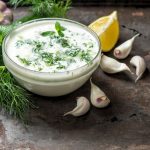 Salsa allo yogurt: la ricetta classica e 2 varianti 1 su tavolo da cucina, ciotola di vetro con la salsa allo yogurt, attorno gli ingredienti limone, erbe aromatiche e aglio