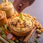 Hummus di ceci fatto in casa: ricetta facile e cremosa 1 su tavolo grigio tagliere con ciotole piene di hummus con attorno ceci sparsi e accanto pinzimonio. Si vede la mano di una donna ch mette nell'hummus un sedano