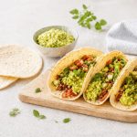 Tortillas di mais: la ricetta per farle morbide in padella 1 Su un piano di marmo bianco c'è un tagliere di legno con delle tortillas alcune sono condite con avocado e dei fagioli. Sullo sfondo c'è una ciotola con della guacamole e un canovaccio bianco.