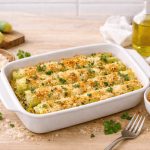 Porri gratinati al forno senza besciamella: ricetta facile e leggera 1 In primo piano su un tavolo di legno chiaro c'è una pirofila di ceramica bianca con dei porri gratinati al forno. Intorno al tavolo c'è del pangrattato in una ciotolina, dell'olio EVO e del parmigiano per la panatura.