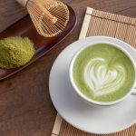 Matcha latte: la bevanda verde che conquista al primo sorso 1 stuoia di bambù con piattino e tazza piena di matcha latte con un disegno a cuore, frusta e polvere matcha accanto