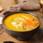 Vellutata di carote e patate: ricetta light e video ricetta 1 In primo piano su un tavolo di legno, in una ciotola scura c'è una vellutata di carote cremosa e sana. Sullo sfondo ci sono delle carote, una cipolla e un tovagliolo chiaro.