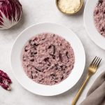 Risotto al radicchio rosso: la ricetta facile e cremosa step by step 1 In primo piano su un tavolo di marmo chiaro c'è un piatto di risotto al radicchio. Sulla destra c'è una forchetta dorata con un tovagliolo color tortora. Sullo sfondo un cespo di radicchio rosso di Treviso.