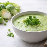 Zuppa di cavoletti di Bruxelles: ricetta passo passo 1 In primo piano su un tavolo di marmo bianco c'è un piatto con la zuppa di cavoletti di Bruxelles condita con delle foglie di prezzemolo. Sullo sfondo alla sinistra ci sono dei cavoletti di Bruxelles.
