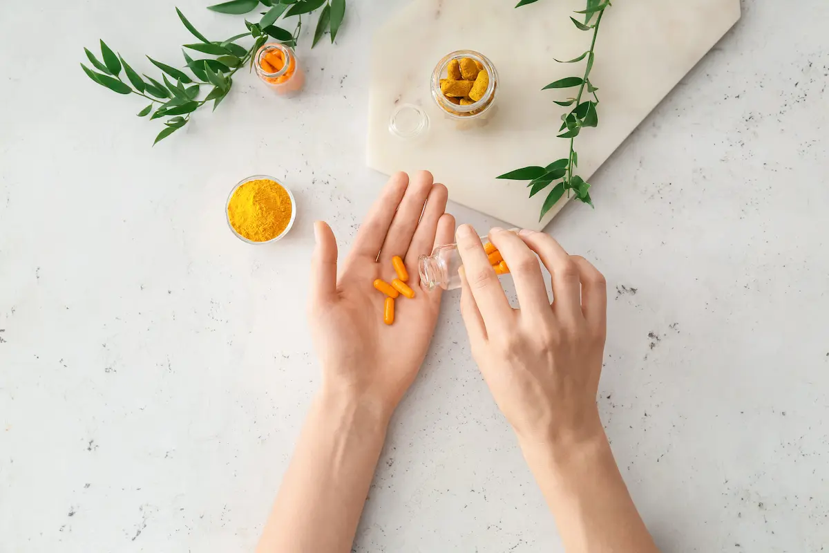 Curcumina: proprietà, benefici e i migliori integratori 2 su tavolo bianco, mani di una donna che versa integratori di curcumina nella mano con accanto la polvere di curcuma