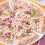 Pizza con Cotechino Modena IGP: la ricetta gustosa 2 su un tavolo con tovaglia gialla, una pizza con cotechino e carciofi tagliata in 8 su un piatto bianco e accanto forchetta e coltello con un tovagliolo bordeaux