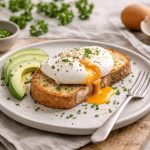 Uova in camicia: ricetta veloce con tuorlo morbido 1 In primo piano su un tavolo di legno c'è un piatto con uova in camicia su una fetta di pane tostato, dell'erba cipollina tritata sopra e delle fettine di avocado.