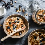 Granola fatta in casa: ricetta step by step e video ricetta 1 In primo piano su una tovaglia scura, due ciotole con della granola fatta in casa, dello yogurt e dei mirtilli. Sulla destra un barattolo in vetro che conserva della granola mentre sulla sinistra c'è un bricchetto con del latte.