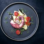Polpo alla griglia: un secondo piatto leggero e gustoso 1 su sfondo nero, piatto nero con polpo alla griglia su un nido di purè di patate