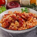 Pasta con pomodori secchi: un primo piatto veloce e saporito 1 piano di cucina con piatto fondo con pasta con pomodori secchi, tovagliolo piegato e una forchetta, e ingredienti della preparazione