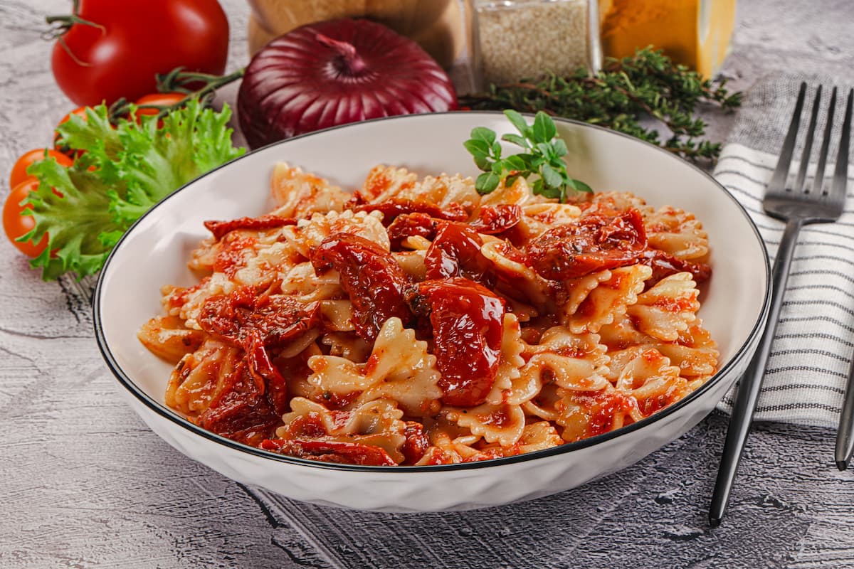 Calorie della pasta: è vero che la pasta fa ingrassare? 1 piano di cucina con piatto fondo con pasta con pomodori secchi, tovagliolo piegato e una forchetta, e ingredienti della preparazione