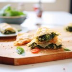 Spanakopita: ricetta tradizionale e varianti gustose 1 su tavolo di legno bianco un tagliere con la Spanakopita tagliata e fuori fuoco un piatto di spinaci freschi