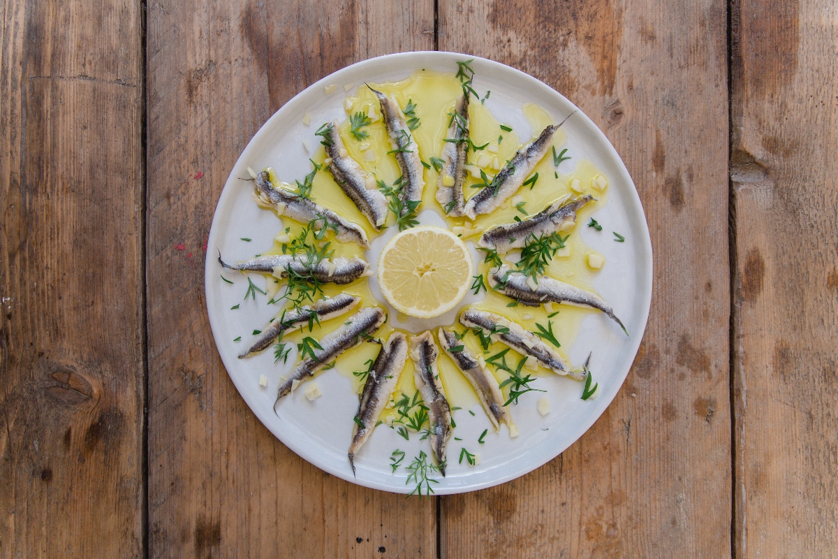 Ricette con alici: 12 idee semplici e ricche di sapore 2 su tavolo di legno, piatto bianco con alici marinate, olio Evo e una fetta di limone al centro