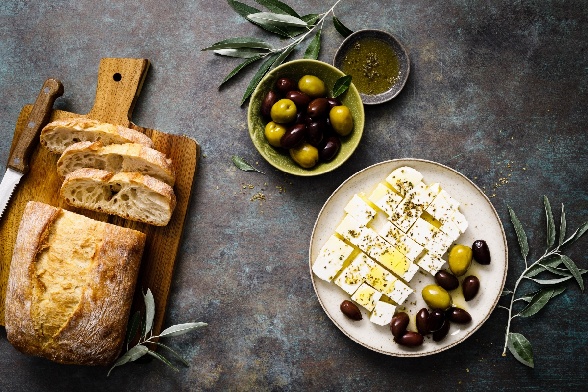su banco grigio di cucina, tagliere con pane tagliato e Coltello con accanto un piatto con feta greca tagliata con olio EVO e olive verdi e nere. Dietro ciotola verde con olive