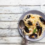 Spaghetti con le cozze: un primo piatto della cucina mediterranea 1 su un tavolo di legno bianco un piatto grigio con gli spaghetti con le cozze una forchetta e un cucchiaio