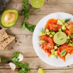 Carpaccio di salmone marinato al limone 1 In primo piano su un tavolo di legno, in un piatto fondo bianco c'è un carpaccio di salmone condito con avocado e lime. Sulla sinistra delle fette integrali con semi, spicchi d'aglio, basilico e una metà di un avocado.