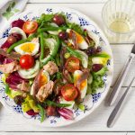 Insalata nizzarda: la ricetta completa con tonno, uova e verdure 1 In primo piano su un tavolo in legno bianco c'è un piatto fondo con dell'insalata nizzarda. Sulla destra ci sono due posate e un bicchiere di vetro.