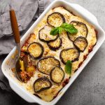 Moussaka: la ricetta greca con besciamella senza burro 1 In primo piano su uno sfondo grigio c'è una pirofila bianca con la moussaka di melanzane.
