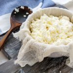 Ricetta fiocchi di latte: come preparare il Cottage Cheese in casa 1 su tagliere di legno ciottola con fiocchi di latte fatto in casa e cucchiaio di legno