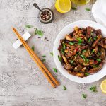 Melanzane in agrodolce alla Siciliana: ricetta originale e la ricetta al forno 1 su tavolo piatto di marmo, piatto bianco con melanzane in agrodolce, prezzemolo, limone e pepe in una ciotola. Accanto bacchette cinesi e limone tagliato
