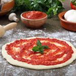 Pizza rossa: consigli e ricetta per una pizza croccante 1 su piano da cucina di legno, pizza rossa cruda con passata di pomodori, un mattarello, cipolla, basilico, una ciotola con 2 mozzarelle