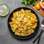 Pasta con pesce spada: un primo piatto della cucina mediterranea 1 su tavolo grigio, piatto nero con pasta con pesce spada, prezzemolo e pomodorini gialli. Accanto una forchetta e un coltello e dietro un tagliere con pomodorini gialli e prezzemolo