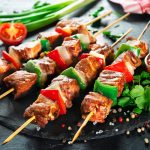 Spiedini di carne: la ricetta e come prepararli 1 su tavolo nero primo piano di un piatto con spiedini di carne e verdure in un piatto nero con pomodorini tagliati e una canovaccio rosso e bianco