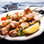 Spiedini di pesce: la ricetta e come cucinarli 2 su tavolo di legno piatto bianco con spiedini di pesce e una fetta di limone, in secondo piano pepe nero