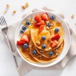 Pancake proteici: ricchi di proteine, sani e sazianti 1 su base bianca una scodella con 4 pancake proteici con sopra fragole, mirtilli e frutta secca