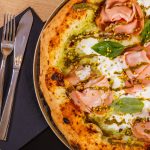 Pizza mortadella e pistacchio: ricetta e segreti di una pizza gourmet 2 primo piano di una pizza mortadella e pistacchi con mozzarella su un tavolo e accanto tovagliolo di carta blu con forchetta e coltello