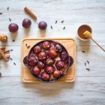 Prugne cotte: come prepararle in modo semplice a casa 1 base di legno con padella piena di prugne cotte, accanto a miele, bastoncini di cannella e prugne fresche