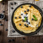 Crepes senza glutine: facili, leggere e deliziose 1 su tavolo di legno, canovaccio marrone con una padella e diverse crepes senza glutine con sopra dei mirtilli e accanto una ciotola piccola con mirtilli