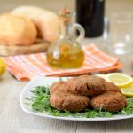 Mondeghili: la ricetta delle tipiche polpette milanesi 2 su tavolo da cucina, piatto con rucola, limoni tagliati e mondeghili, dietro una bottiglietta di olio EVO e panini