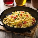 Spaghetti aglio olio e peperoncino: un primo piatto salva cena 1 Su tavolo di legno, tagliere, una padella con Spaghetti aglio, olio e peperoncino , dietro una grattugia e un bicchiere di olio d'olive