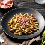 Pasta con Mortadella Bologna IGP e zucchine 2 su tavolo di legno scuro, piatto nero con dentro pasta con mortadella e zucchine, accanto un piattino con mortadella e in basso a destra 2 fiori di zucchina accanto alla forchetta