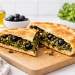 Pizza di scarola napoletana: ricetta originale step by step 1 In primo piano su un tagliere di legno chiaro una pizza di scarola con olive e capperi e acciughe. Sullo sfondo ci sono un'oliera, una ciotola bianca con delle olive nere e la scarola.