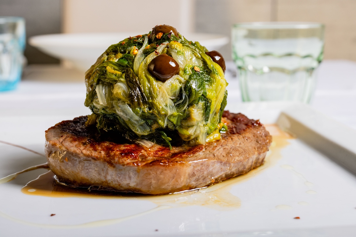 Le ricette con la scarola: dalle classiche napoletane alle varianti 5 su tavolo bianco: una fetta di carne di manzo con scarola in padella sopra a forme di palle con olive nere di Gaeta
