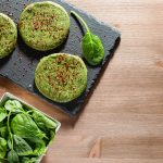 Burger di spinaci fatti in casa: la ricetta veloce e gustosa 1 su tavolo di legno sotto piatto di ardesia con 3 burger di spinaci, un piccolo contenitore di peperoncino e a destra contenitore con spinaci freschi