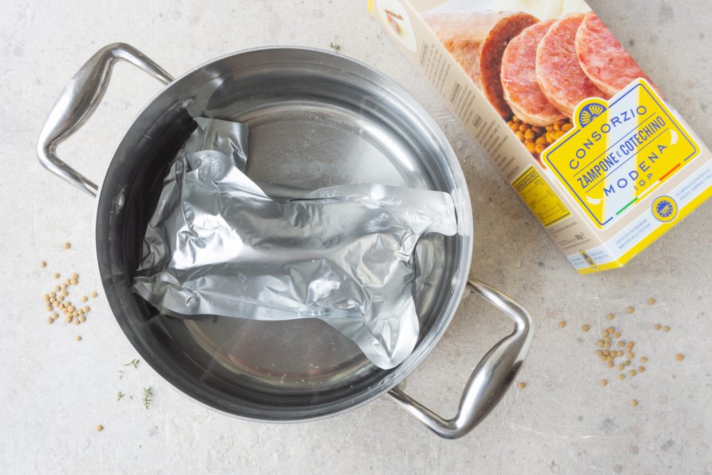 Cotechino Modena IGP e lenticchie, la ricetta classica passo passo e video ricetta 6 pentola di alluminio con acqua e confezione di cotechino sottovuoto immersa dentro, a destra pacco della confezione
