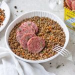 Cotechino Modena IGP e lenticchie, la ricetta classica passo passo e video ricetta 2 su tavolo con tovaglia bianca, insalatiera di ceramica bianca con cotechino e lenticchie , sulla destra piatto pieno della pietanza con forchetta e dietro confezione della scatola del cotechino Modena IGP
