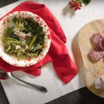Minestra maritata con Salamini Italiani alla Cacciatora DOP 2 su tavolo nero tagliere bianco con un contenitore con minestra maritata con salamini sulla destra; tagliere di legno con un salame cacciatore e sulla sinistra 2 rami di legno con fiocco rosso