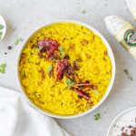 Risotto allo zafferano con Salamini Italiani alla Cacciatora DOP 2 su piano di lavoro bianco, un piatto che contiene riso allo zafferano con salamini sulla destra 2 Salamini Italiani alla Cacciatora DOP , a sinistra erbe aromatiche e sotto a sinistra piatto con zafferano