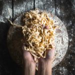 Tagliatelle fatte in casa: la ricetta con consigli su condimenti e cottura 1 Su piano di lavoro di legno scuro, 2 mani che prendono tagliatelle fatte in casa. Sotto tagliere di legno pieno di farina