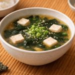 Zuppa di miso: la ricetta giapponese con foto passo passo 1 In primo piano una zuppa di miso con cubetti di tofu, alghe wakame e cipollotto verde in una ciotola bianca su una tovaglietta in legno.