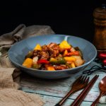 Maiale in agrodolce, la ricetta cinese da fare in casa 1 su tavolo di legno azzurro, piatto blu con maiale in agrodolce, una ricetta cinese con tovagliolo beige e accanto fiori bianchi, dall'altra parte poste, peperoncino e macinapepe di legno
