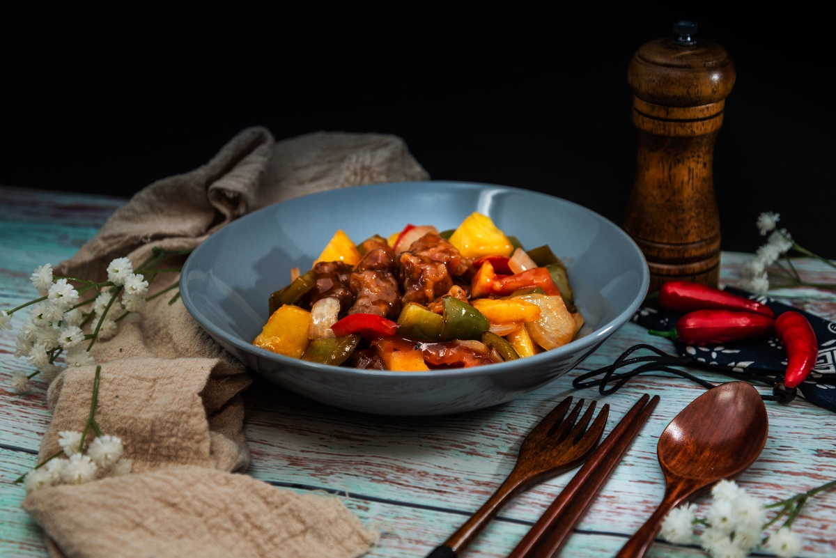 Ricette cinesi facili: antipasti, primi e secondi della cucina cinese 3 su tavolo di legno azzurro, piatto blu con maiale in agrodolce, una ricetta cinese con tovagliolo beige e accanto fiori bianchi, dall'altra parte poste, peperoncino e macinapepe di legno