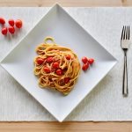 Pasta con pomodorini: un primo piatto veloce e gustoso 1 su tavolo di legno sottopiatto bianco di stoffa con un un piatto quadrato pieno di pasta con pomodorini freschi. Accanto al piatto 5 pomodorini e a destra forchette e coltello